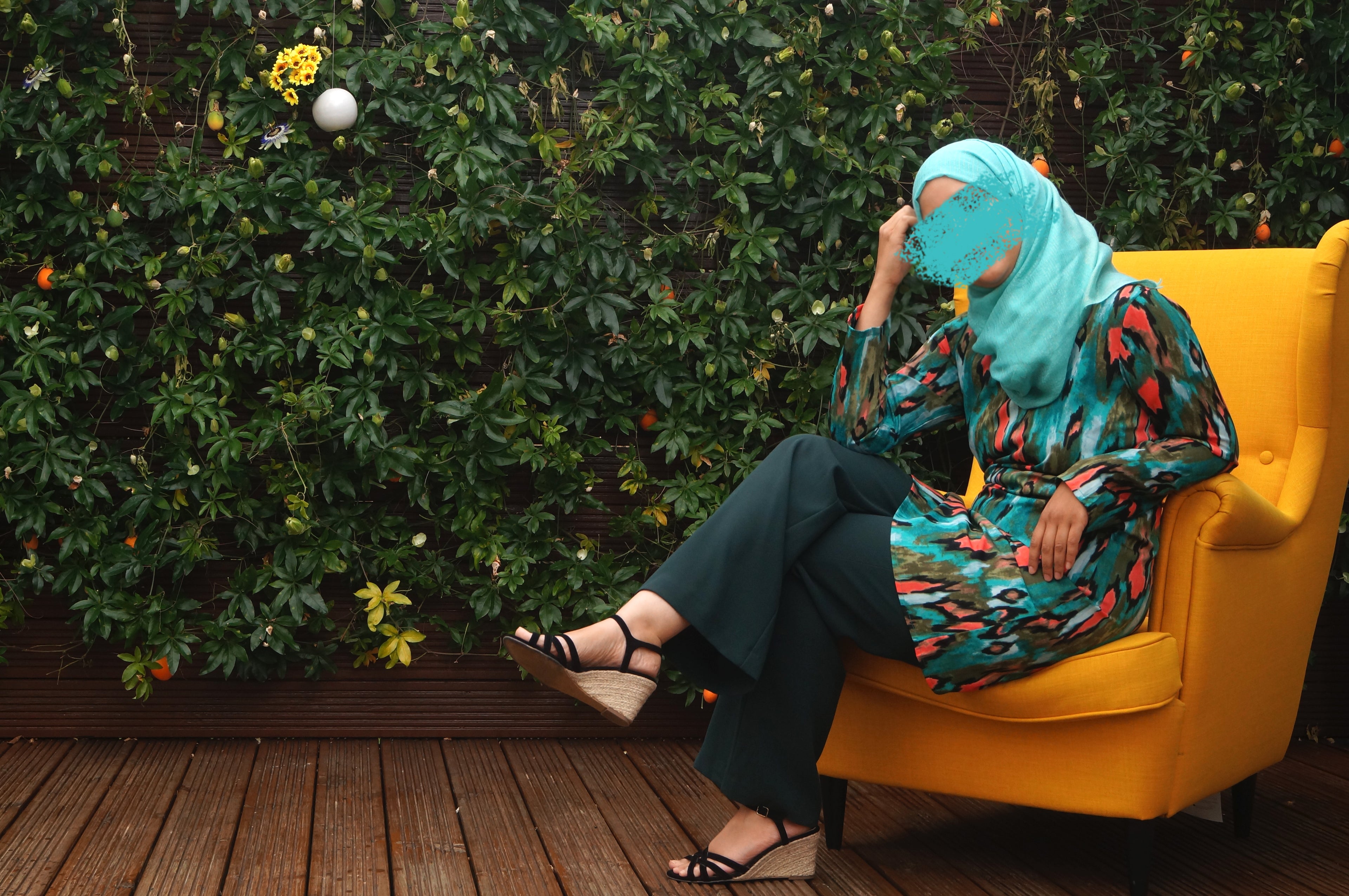 Person wearing a colorful outfit and blue headscarf sitting on a yellow chair with green foliage in the background.